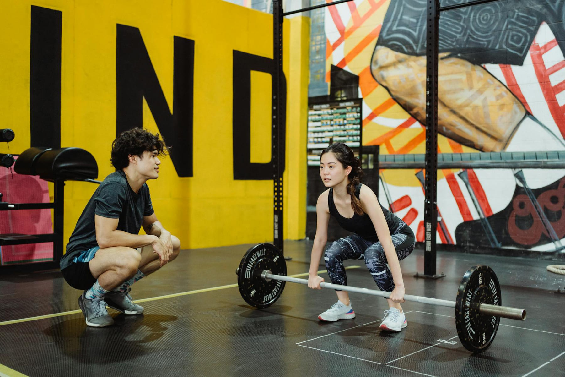 A young woman lifting weights guided by a trainer in a colorful gym setting. on First Rep Forward
